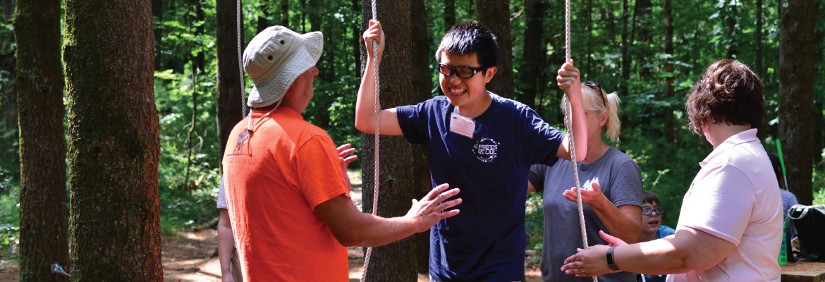 A teenager wearing glasses smiles as he holds two vertical ropes in an outdoor setting.