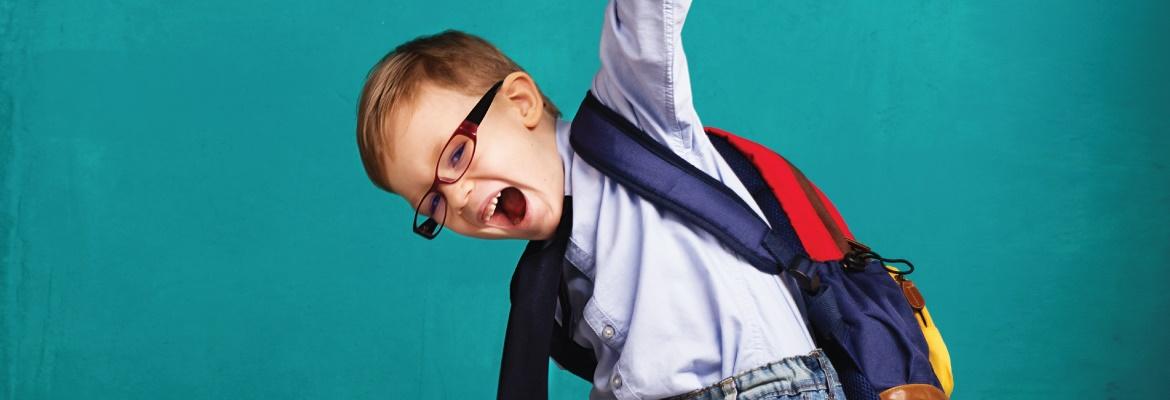 A young child wearing glasses and a backpack smiles while raising a triumphant fist in the air.