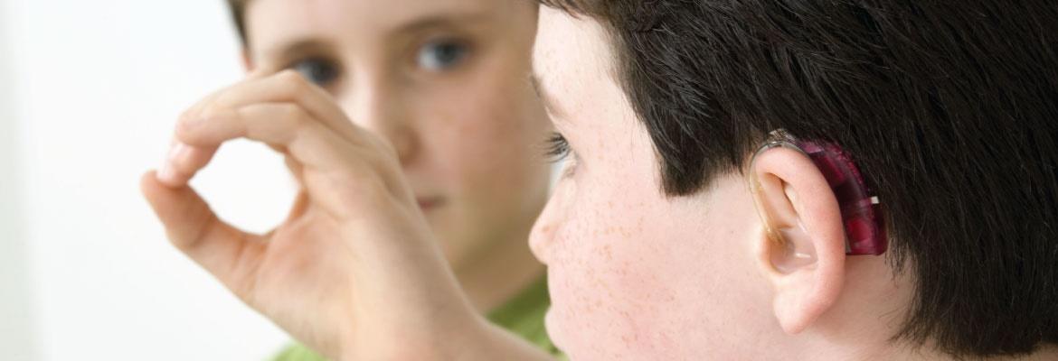 View from behind a young boy who is wearing a hearing device. Another young boy in front of him signs the letter