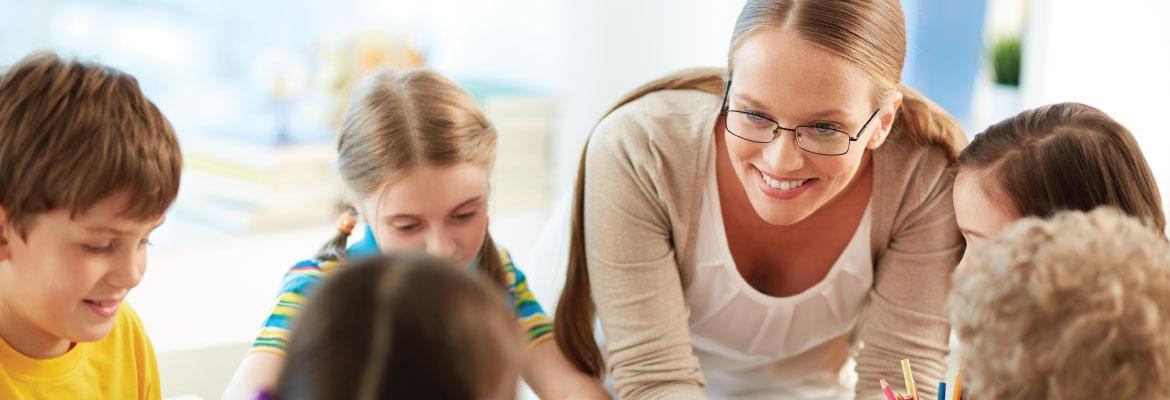 A female teacher wearing glasses leans over and smiles at a child who is seated with four other children at a table.