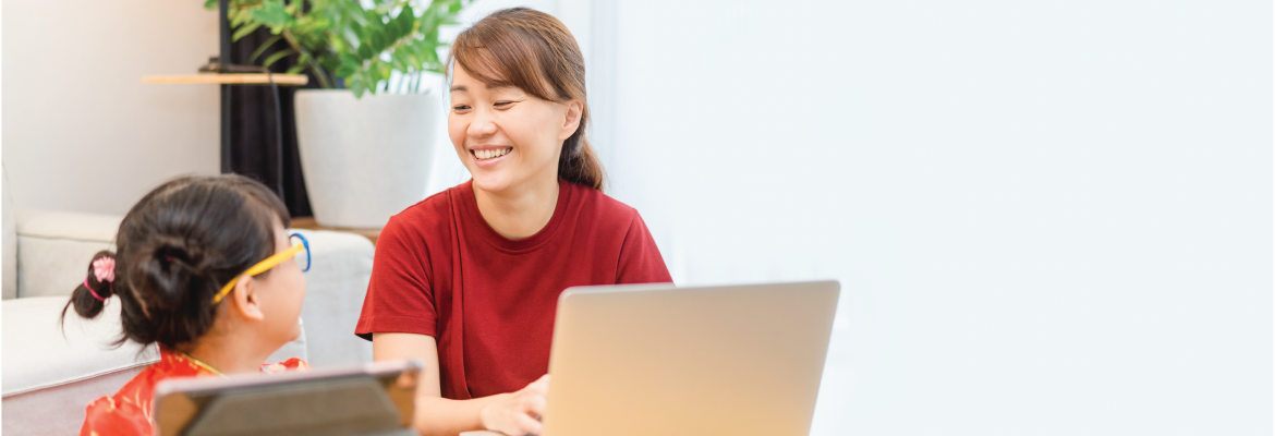 A mother and daughter smile while sitting at a table with a laptop and tablet.