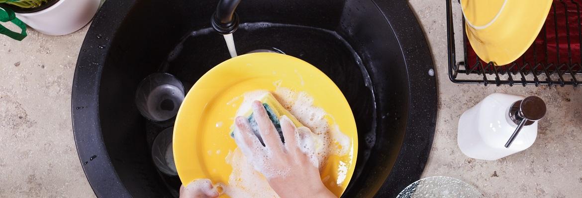 Closeup of a child's hands washing a yellow plate with a sponge over a black sink basin.