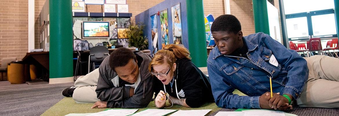 Three students, two boys and one girl, complete a worksheet together while lounging on a carpeted floor at a Finance Park facility.