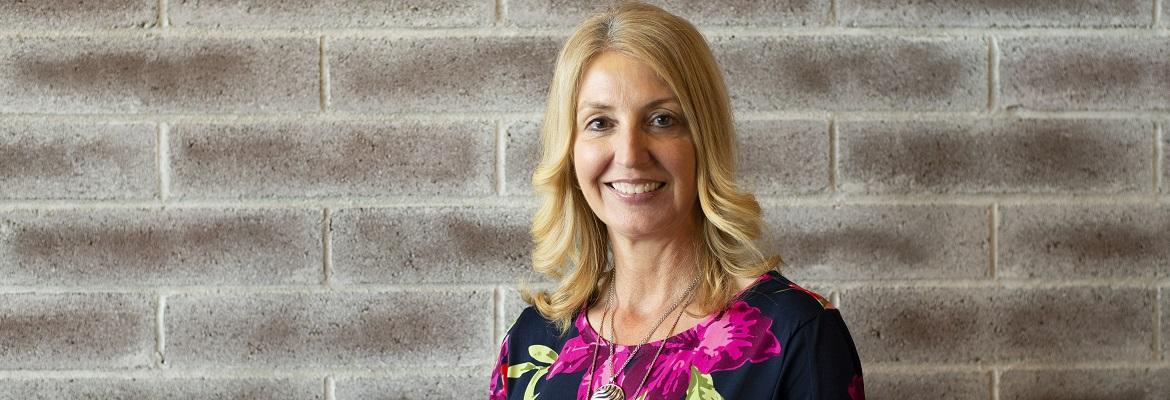 Lynn Pensari smiles for a photo against a distressed brick wall. She has shoulder-length blonde hair and is wearing a pink floral top.