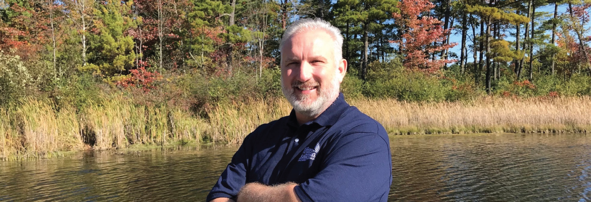 Crossing his arms, Matt Polzin smiles at the camera while standing next to a lake in a wooded area.