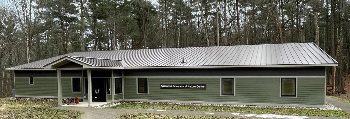 The Sassafras Science and Nature Center in a clearing in the woods. The rectangular building is painted sage green on the bottom and dark brown at the top.