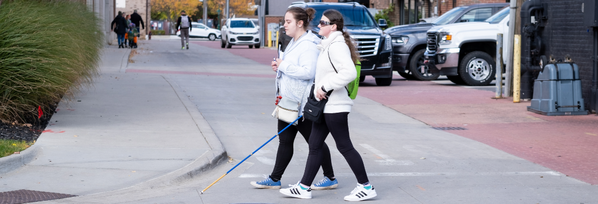 A girl with a white cane and a friend walk across a city street. 