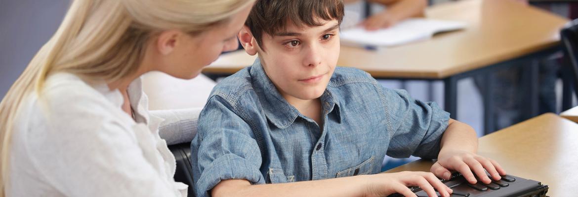 A teacher sits at a desk next to a boy who is using a small rectangular assistive technology device.