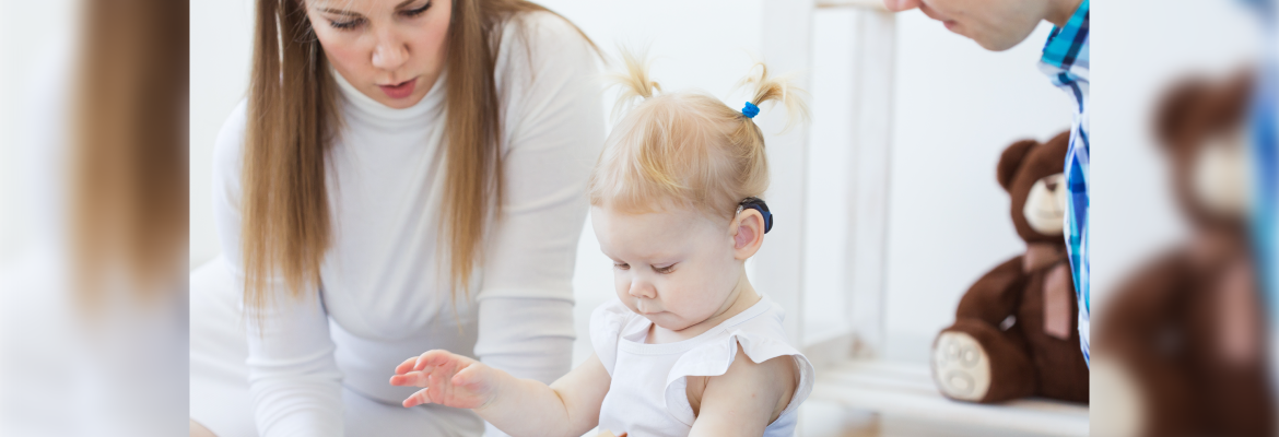 A young girl wearing a hearing aid at home. Two adults are sitting by her. 