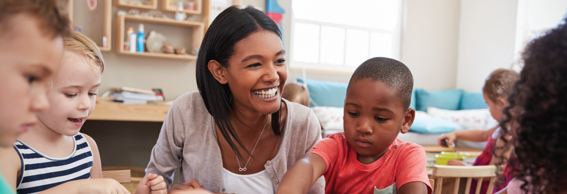 A teacher smiles while working with a group of children in a classroom.