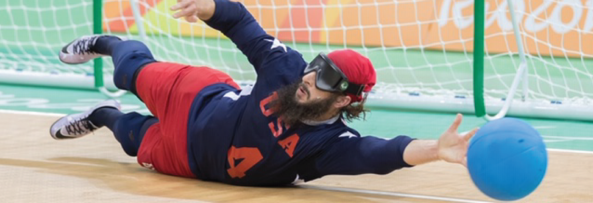 Goalball player John Kusku, wearing an eye cover, slides sideways and stretches his arm out to block a goal.