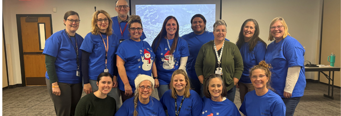 A group of fourteen teacher consultants pose for a group photo in a large room, mostly wearing matching blue shirts.