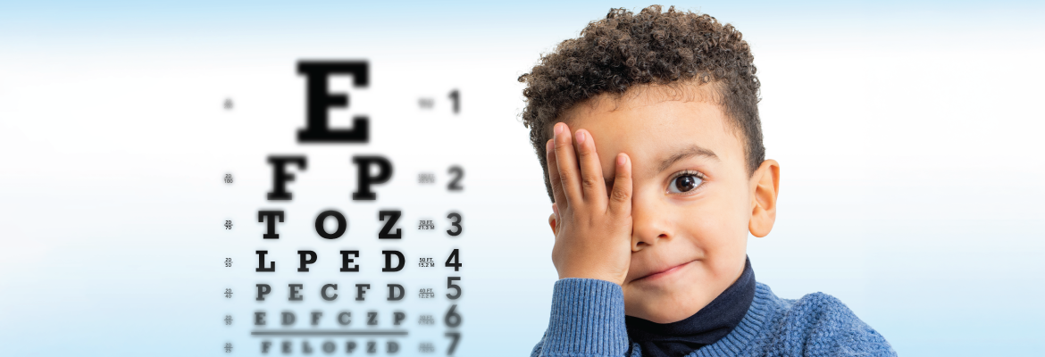 Young child in a blue sweater covering one eye, next to an eye chart during an eye exam.