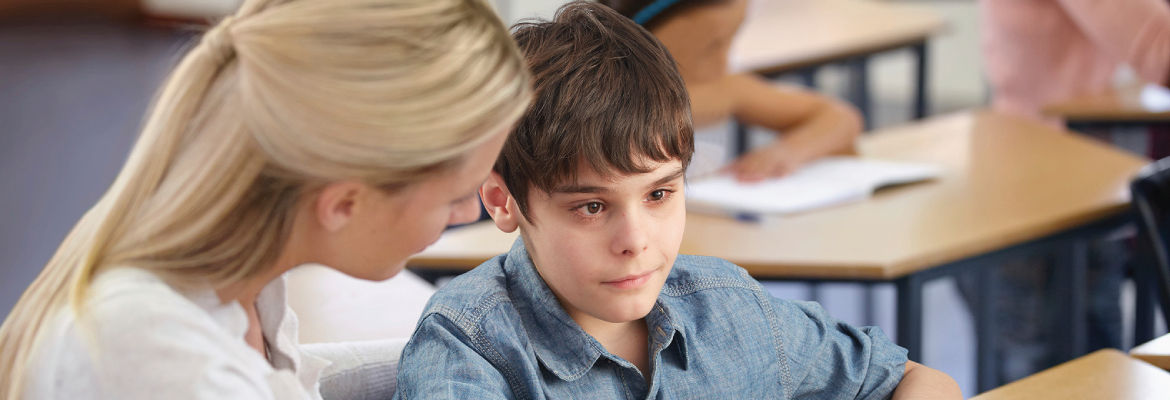 A teacher sits at a desk next to a boy who is looking off-camera.