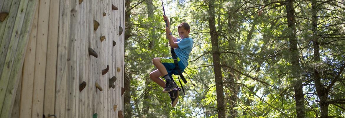 A boy smiles while rappelling down a wooden climbing wall in the woods.