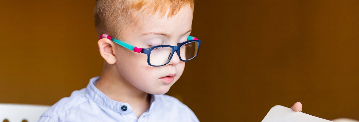A young child with short hair reads a book while wearing colorful blue and pink glasses.