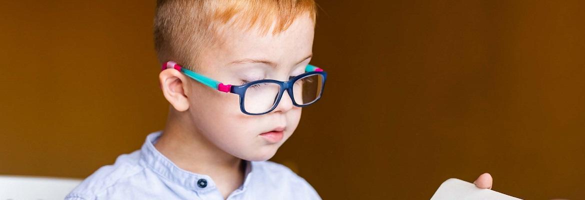 A young boy wearing glasses with a multicolored frame looks down at a book.