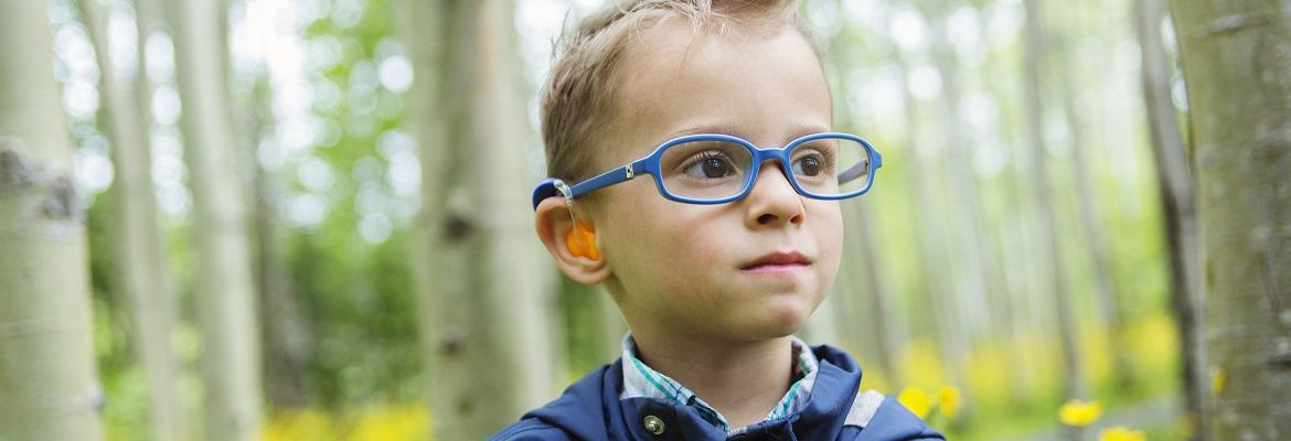 A boy wearing glasses and a hearing device smiles off-camera while standing in a wooded area.