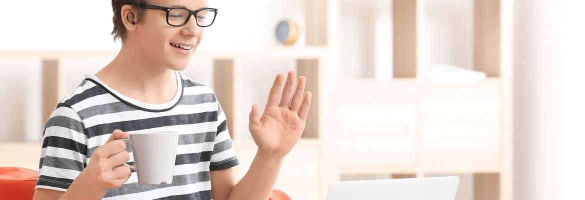 A teenager wearing a hearing device smiles and waves during a video call while holding a mug.
