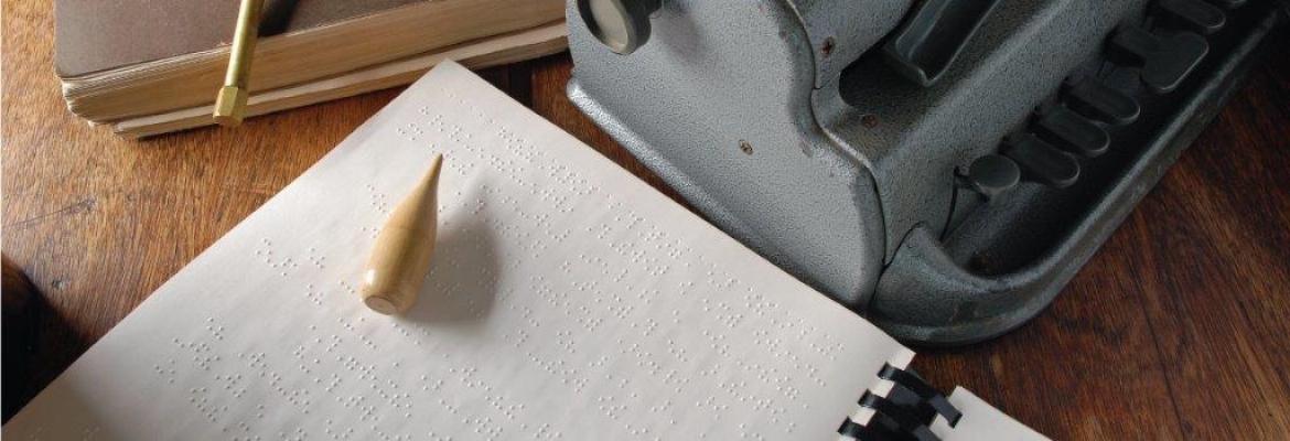 A braille writer rests on a table next to a sheet of braille and other braille tools.