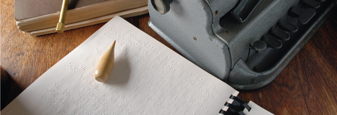 A braille writer sits on a wooden table next to a sheet of embossed braille.