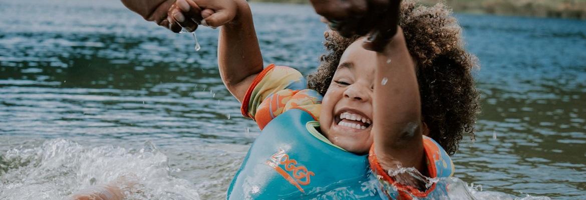 A young child laughs while holding an adult's hands and playing in a lake.