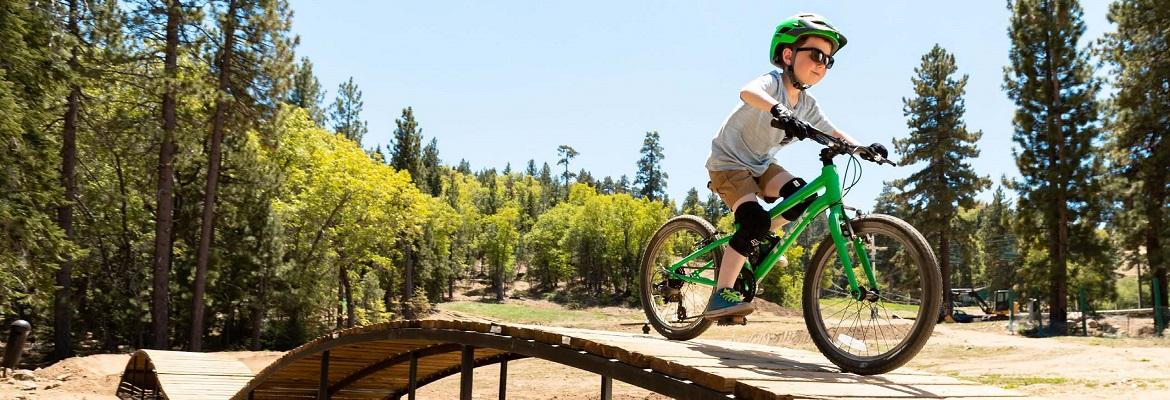 A young child smiles while riding a green bike on a wooden ramp outside.