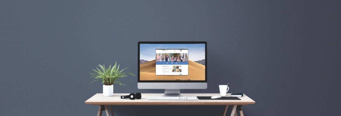 A large computer monitor on a desk, showing the MDE-LIO website. The desk features a plant, headphones, keyboard, and mug.