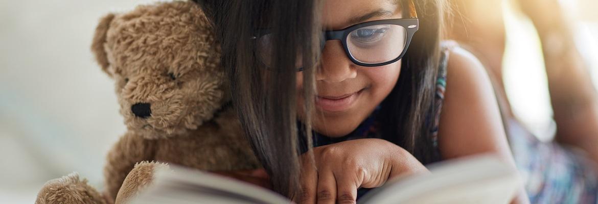 A young girl smiles while holding a teddy bear and reading a book