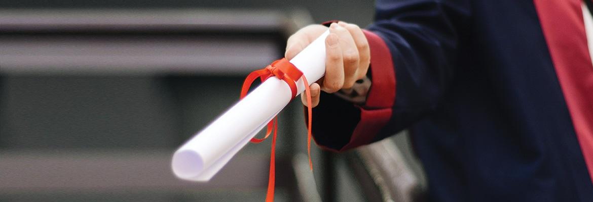 A student in a black robe holding a diploma that is rolled up and tied with red ribbon.