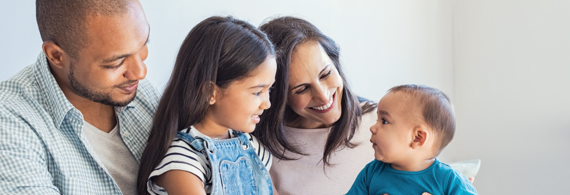 A mother, father, and child smile at a baby the mother is holding.