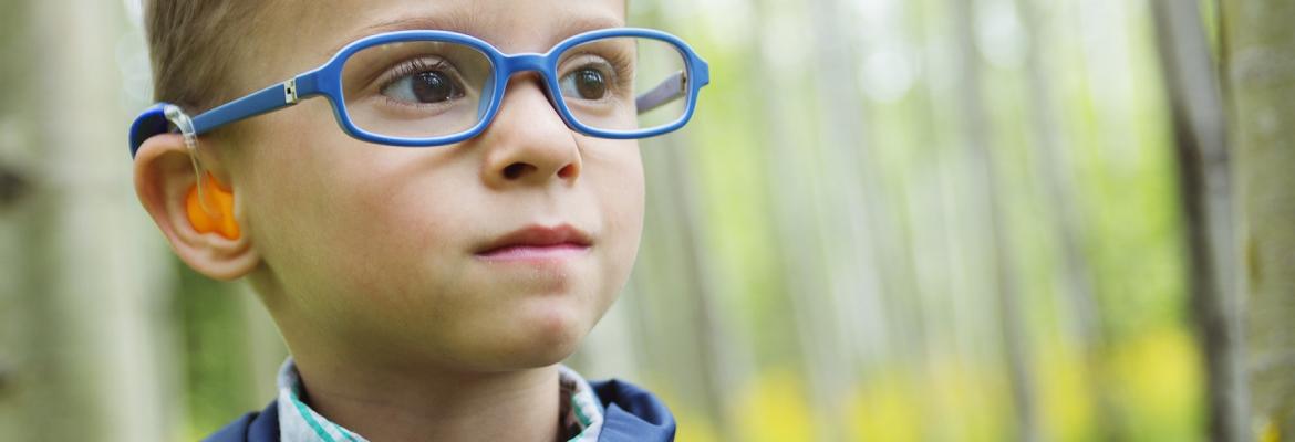 A young boy wearing glasses and a hearing device smiles while standing outside.