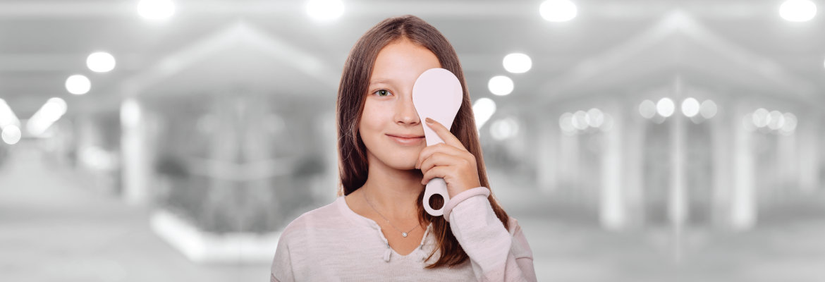 A teen girl smiles at the camera while covering one eye with a pink paddle.