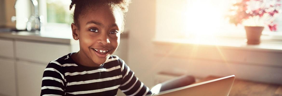 A young girl smiles at the camera while sitting in front of a laptop at home.
