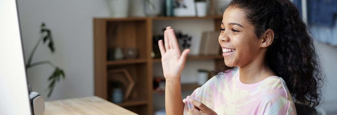 A pre-teen girl wearing a ponytail smiles and waves while on a video call.
