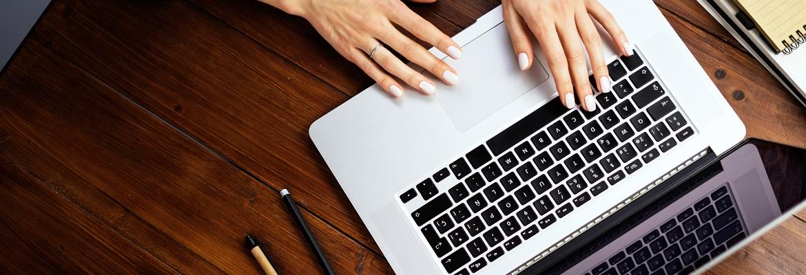 Closeup of hands resting on a silver laptop that is sitting on a brown wooden table.