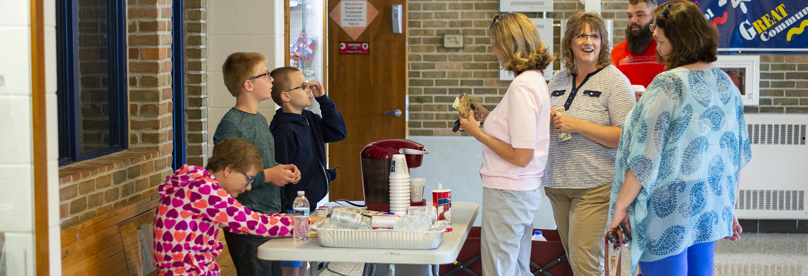 One girl sits and two boys stand at a folding table that features a single-cup coffee machine, cups, and bottles of water. One of the boys talks to a woman who is in line to buy coffee on the other side of the table.
