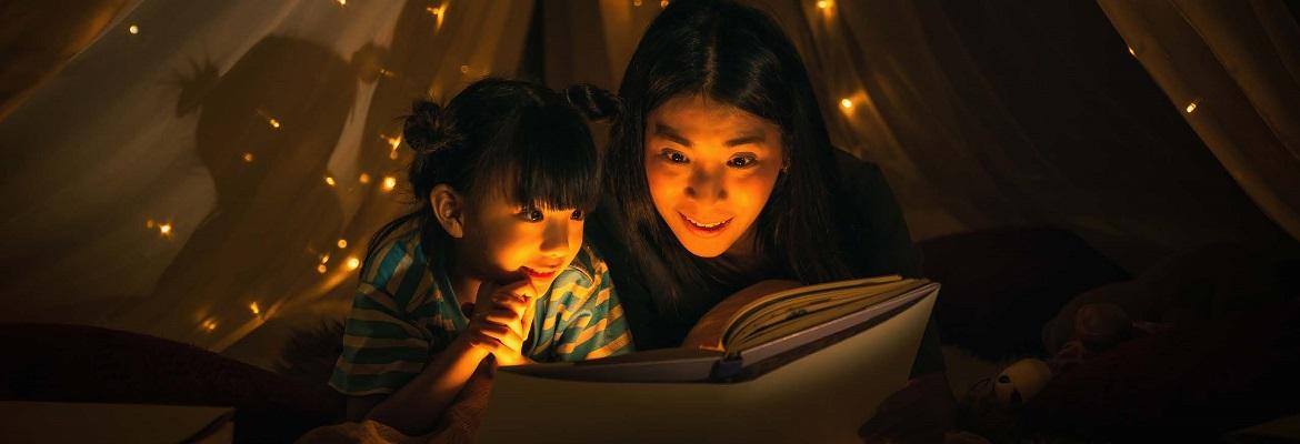 A woman and young daughter smile while reading a book in a tent at night, surrounded by twinkle lights.