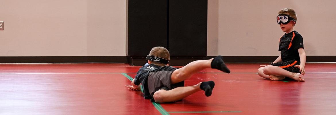 A boy wearing eyeshades sits on the floor at Oak Recreation Center while another student wearing eyeshades dives across the floor to catch a goalball.