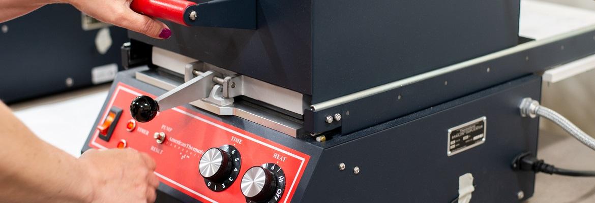 Closeup of a woman's hands pressing the top of a thermoform machine toward the base. A small red instrument panel on the front features a switch and knobs for time and heat.