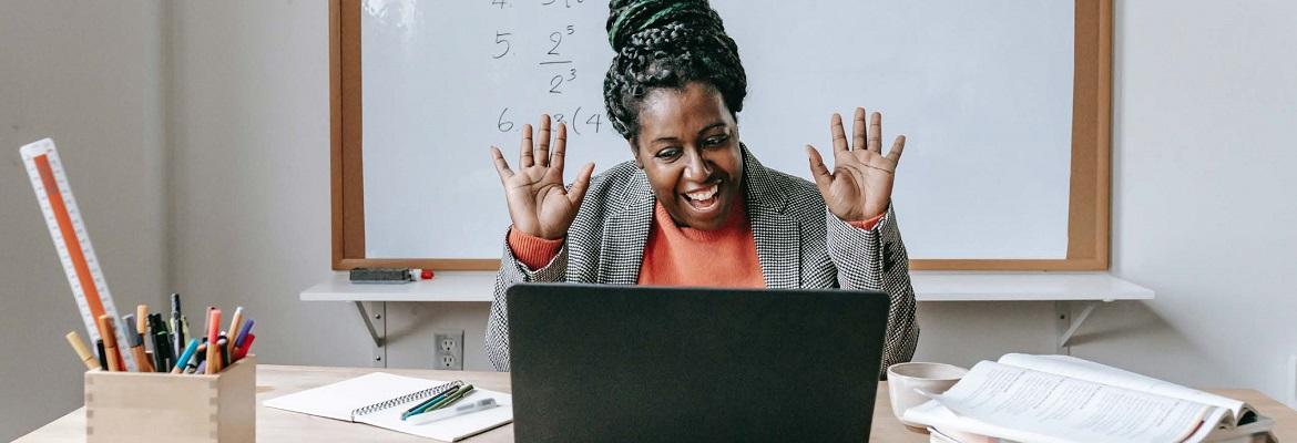 A teacher who is sitting in a classroom smiles and waves at an open laptop in front of her.