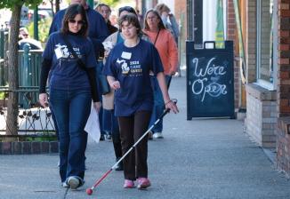 A student and volunteer walk down a busy city street wearing Cane Quest T-Shirts