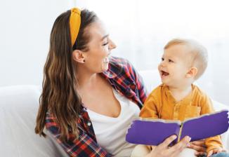 A mother sits on a couch with her toddler in her lap. The both smile as the mother reads a book. 