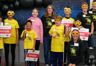 A group of contestants and volunteers at the Michigan Abacus Bee against a starry sky backdrop and black balloons.