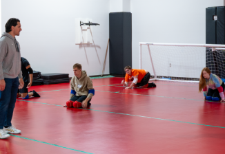 An instructor teaches a group of students how to play goal ball in a gym.