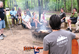 A group of people gathered around a campfire, enjoying music and conversation in a wooded outdoor setting.