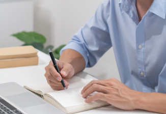 Person writing in a notebook at a desk with a laptop and stacked books in a bright, minimal office setting.
