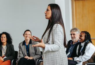 A speaker presents in front of an audience seated in a meeting room, engaged in discussion and learning. A whiteboard is visible.