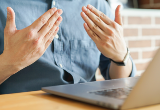 A man sits in front of a laptop at home and uses his hands to communicate.