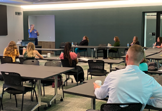 Classroom setting with attendees seated at tables listening to a presenter.